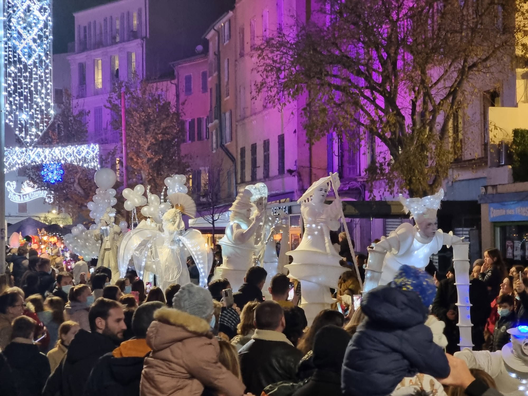 Parade de rue illuminée dans un centre-ville décoré pour Noël, devant une foule dense