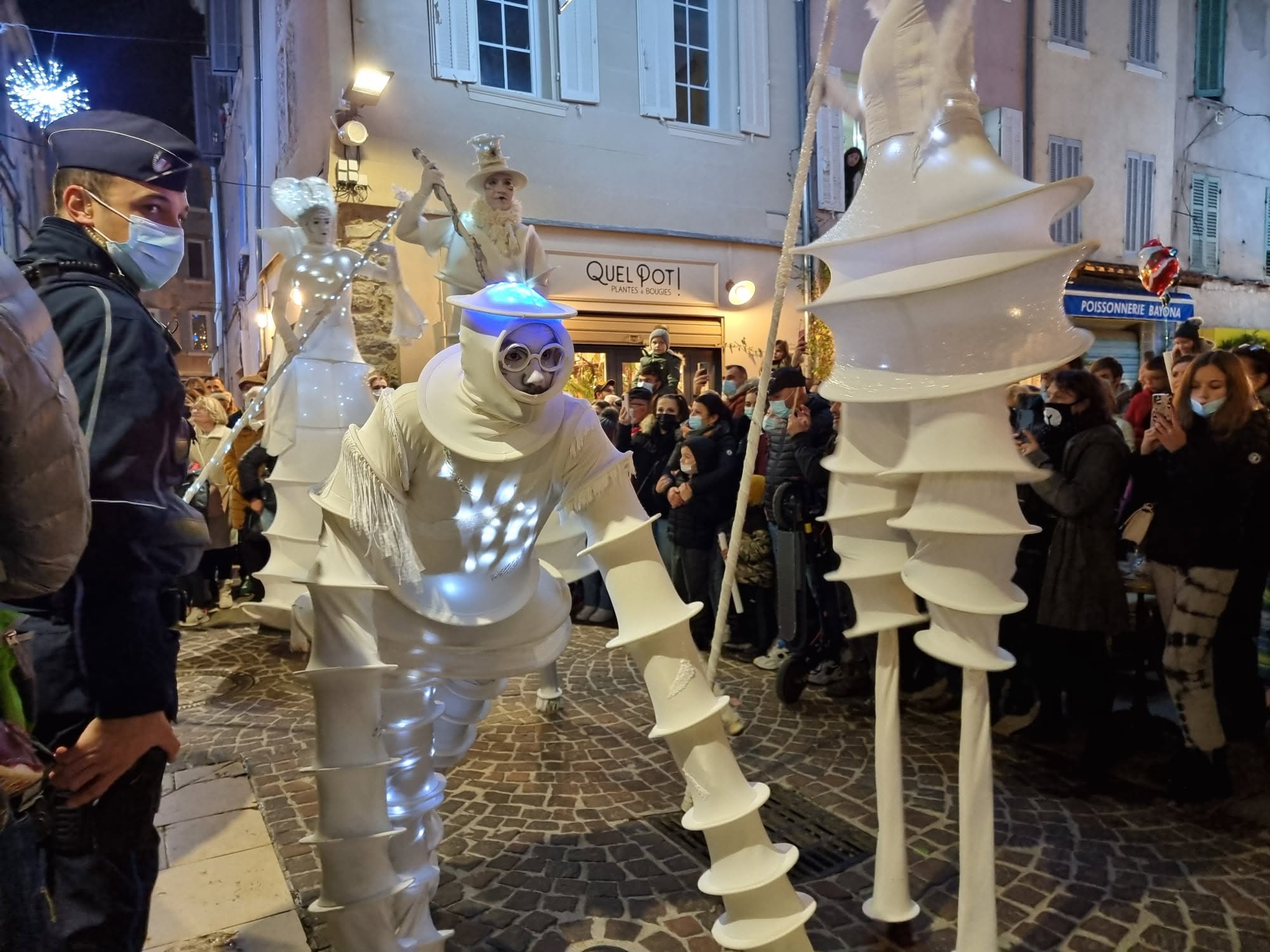 Échassiers en costumes blancs lumineux traversant une rue commerçante bordée de spectateurs