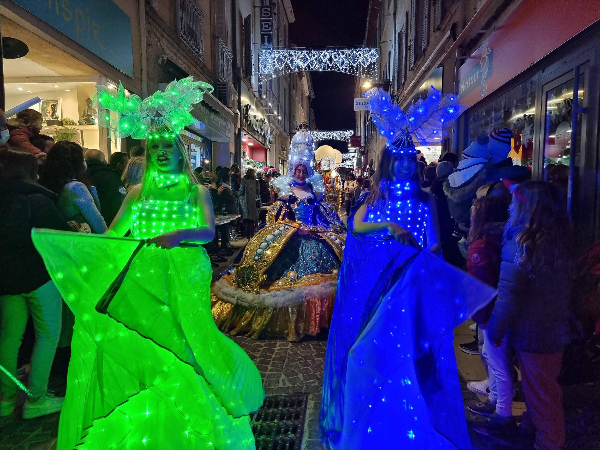 Échassières en costumes LED colorés dans une rue commerçante décorée pour les fêtes
